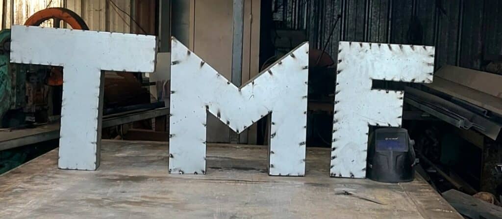 Lettres géantes TMF en acier avec points de soudure apparents dans un atelier de métallurgie.