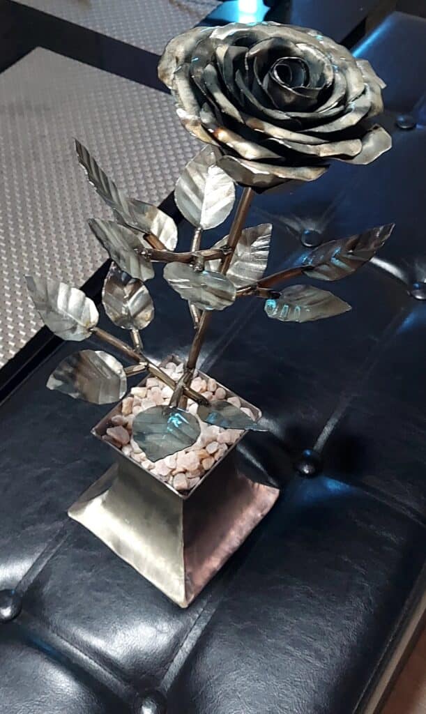 Rose en Métal Artisanal : Sculpture Florale Décorative Sculpture de rose en métal argenté avec feuilles dentelées dans un pot carré rempli de petits cailloux blancs.